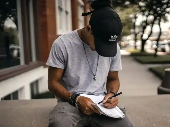 A young man wearing a cap, sitting outdoors and writing in a notebook.