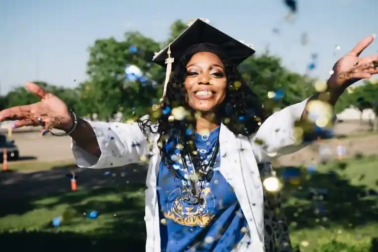 A joyful female graduate wearing a cap and gown celebrating outdoors with confetti.