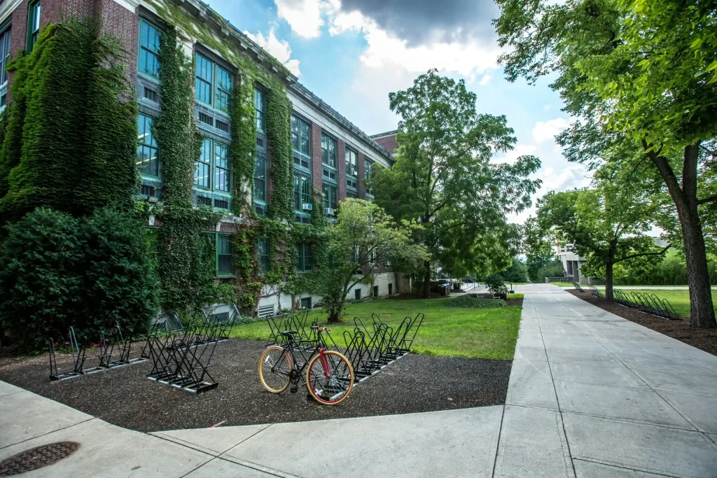 A picturesque campus scene with an ivy-covered brick building, bike racks, and lush green trees under a partly cloudy sky.